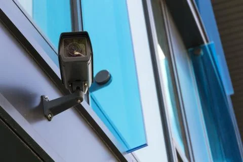 Grey security camera attached to wall blue windows in background Stock Photos
