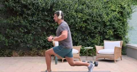 Grey-shirted man following headset beep, jogging doing lunges on patio mat for Stock Footage 345123994