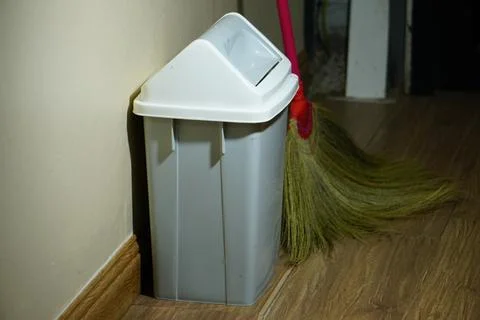 A grey trash bin with a white swing lid stands against a plain cream wall. Stock Photos
