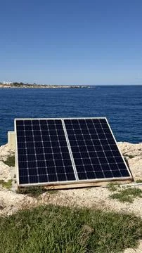 Off-grid solar panel installation on rocky shore with blue ocean and white Stock Photos