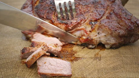 Grilled beef is cut into slices with a knife and fork on a wooden cutting board. Stock Footage 265560921