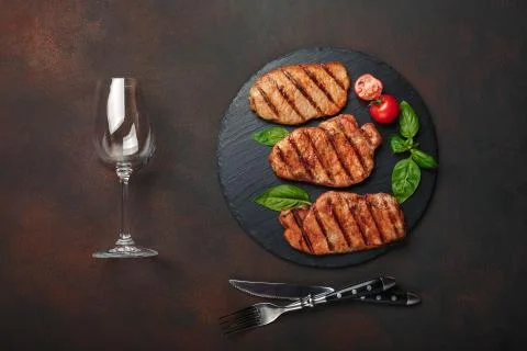 Grilled pork steaks with basil, tomatoes, knife, fork and wine glass on black Stock Photos