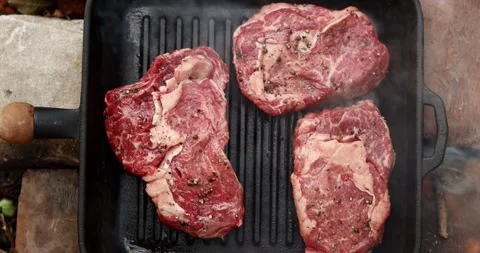 Grilling thick raw beef steaks seasoned with pepper on a cast iron pan Stock Footage 339074415
