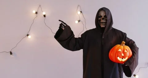 Grim Reaper Pointing Finger Left and holding Pumpkin on Grey Background Stock Footage 313334688