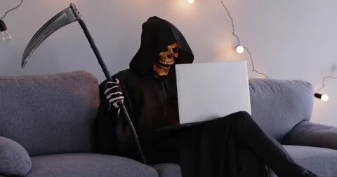 Grim reaper sitting on sofa with scythe in his hand, using laptop computer and Video stock 287410015