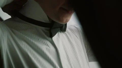 Groom in with a beard stands by the window in a white shirt and examines a bow Stock Footage 140434828