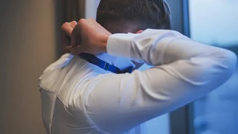Groom getting ready for wedding, adjusting his blue bow tie in front of a window Stock Photos