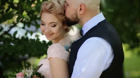 Groom kisses bride's forehead while she leans to him smiling, outdoors Stock Footage 65502004