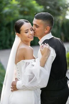 Groom tenderly embracing bride while holding hands in park during wedding c.. Stock Photos