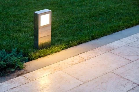 Ground lantern lighting marble walkway in the evening park with a green lawn. Stock Photos