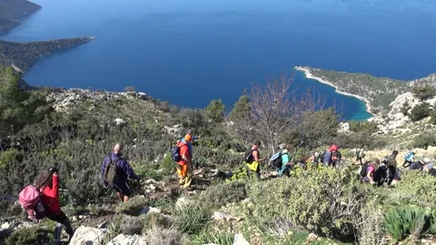 Group of backpackers hiking down a rocky mountain trail overlooking a blue s Vidéo 333464636