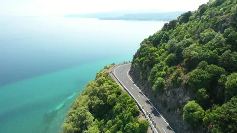 Group of Cyclists ride beatiful curved road above pure Blue Lake - Cinematic Stock Footage 130296279