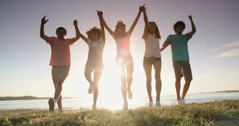 Group of five multiethnic friends running down meadow near lake, jumping Video stock 96427756