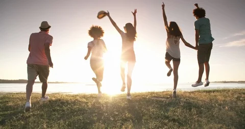 Group of five multiethnic friends running down meadow near lake, jumping Stock-Footage 96428824