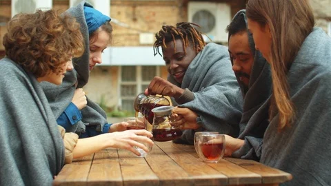 Group of friends in blankets sit outside the cafe and pour tea in cups Stock Footage 121936057