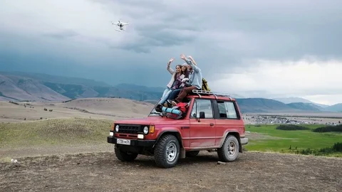 Group of friends doing selfie on drone sitting on red car on mountain backdrop Stock Footage 79734189