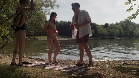 A group of friends sit down on a blanket next to a forest lake holding drinks 스톡 동영상 116759852