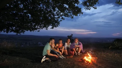 Group of friends sit next to a campfire with warm drinks and talk Stock Footage