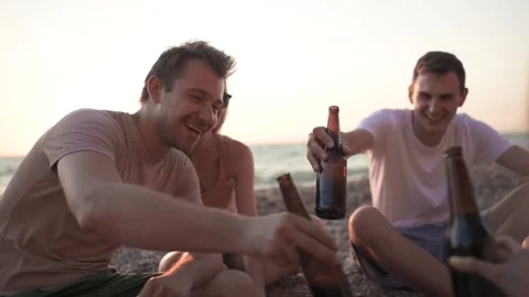 Group of the friends is sitting on the beach, drinking beer and clinking bottles Stock Footage 135257327