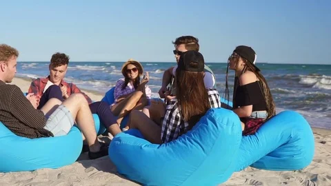 Group of friends sitting on easychairs on the beach playing guitar and singing Stock Footage
