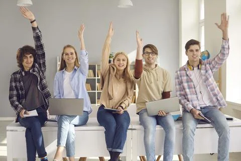Group of happy active school or college students sitting on desk and raising Fotos de archivo