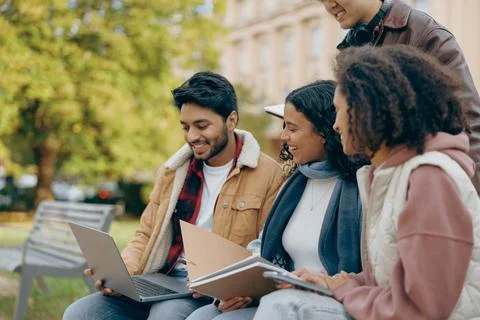 Group of happy students sitting on bench, looking at laptop and discussing their Foto stock