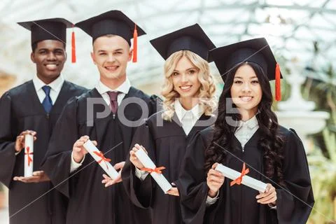 Group of multiethnic students with diplomas in graduation hats standing in ro Stock Photos