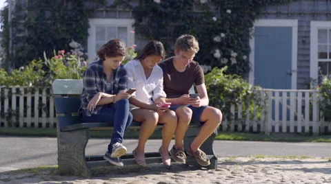 Group Of Multiethnic Teens Sit On Town Bench And Use Their Smart Phones Stock Footage