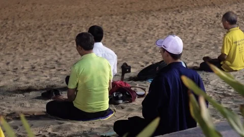 A group of people meditates sitting on the sand at the night beach. Video stock 100876497