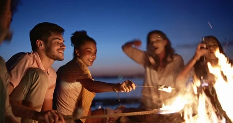 Group of six old multi-ethnic friends sitting near bonfire, roasting sausages Video stock 96454970