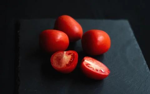 Group of tomatoes, one cut in half, rests on a black board Stock Photos