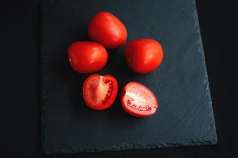 Group of tomatoes, one cut in half, rests on a black board Stock Photos