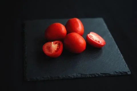 Group of tomatoes, one cut in half, rests on a black board Stock Photos