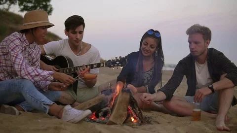 Group of young and cheerful people sitting by the fire on the beach in the Stock Footage
