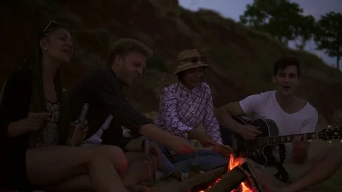 Group of young and cheerful people sitting by the fire on the beach in the Stock Footage