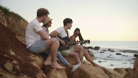 Group of young hipster friends sitting on the rocks by the seashore and playing Stock Footage