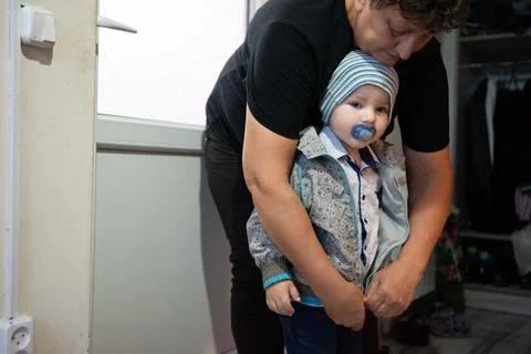 A guardian fastens a patterned jacket on a child with a pacifier and blue s.. Stock Photos
