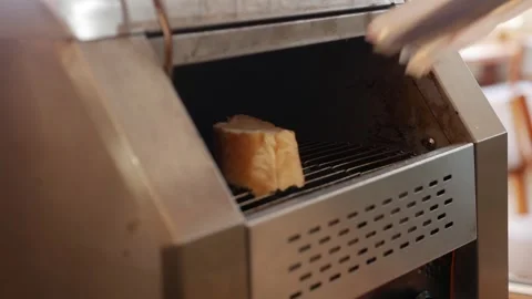 A guest places a slice of bread with metal tongs onto a moving conveyor toaster Stock Footage 332300811