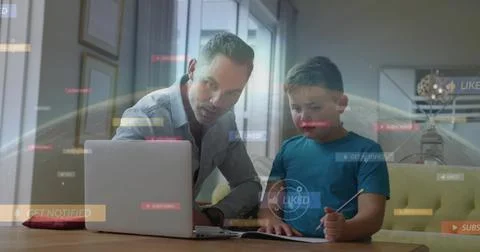 Guiding dad in button-down leaning toward laptop, son in blue tee writing at Stock Photos