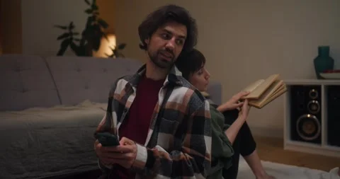 Guy and a girl sit back to back talking while the guy looks at his phone the Stock Footage 318079707