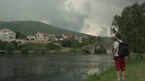 A guy with a backpack approaches the river looking to a beautiful bridge Stock Footage 242918585
