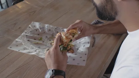 A guy with a beard bites a hamburger sitting at a table in a cafe. man eating Stock Footage 115634249