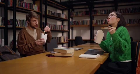 A guy with a beard communicates with an Asian girl while eating his lunch in the Stock Footage 313356687