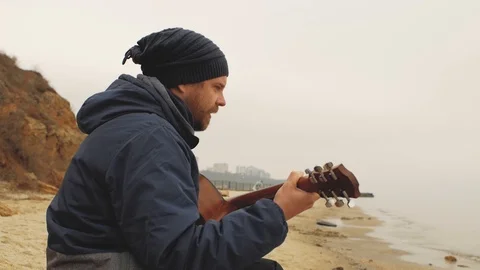 A guy with a beard playing a guitar sitting on a stone by the sea Stock Footage 103445322