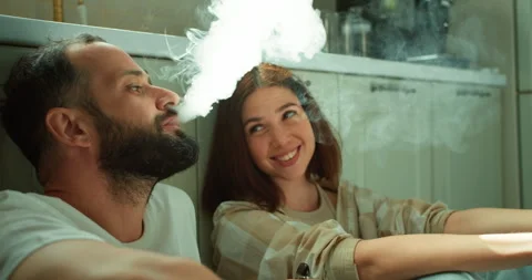 A guy with a beard in a white T-shirt smokes an electronic cigarette with his Video stock 280250894