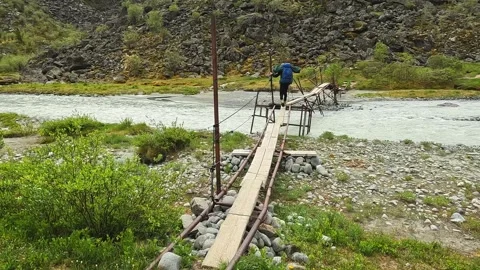 Guy with a big backpack crosses a stormy mountain river on a wooden bridge Video stock 246292177