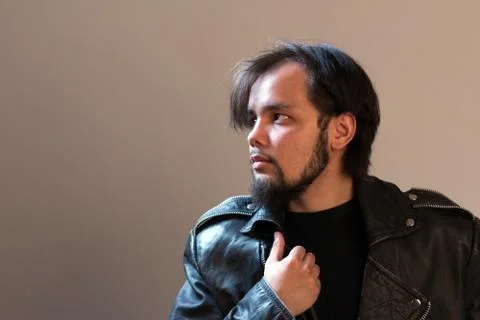 A guy in a black leather jacket with a beard looks away. Beige background Stock Photos