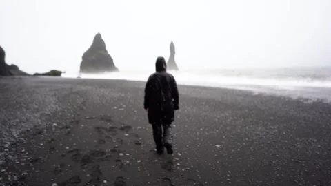 A guy in a black raincoat walks in the rain and storm along a black beach Stock Footage 256887337