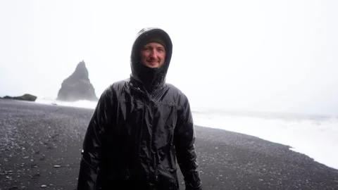 A guy in a black raincoat walks in the rain and storm along a black beach Stock Footage 256887366