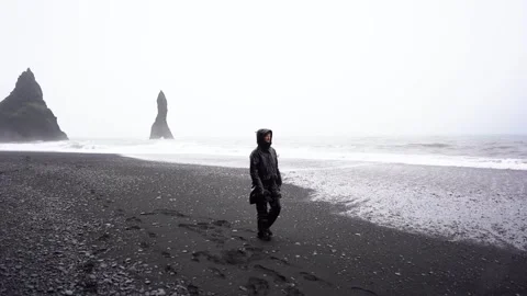 A guy in a black raincoat walks in the rain and storm along a black beach Stock Footage 256887389